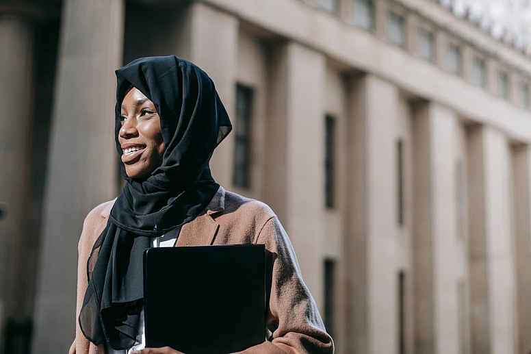 cheerful educated muslim woman with laptop standing outside modern building